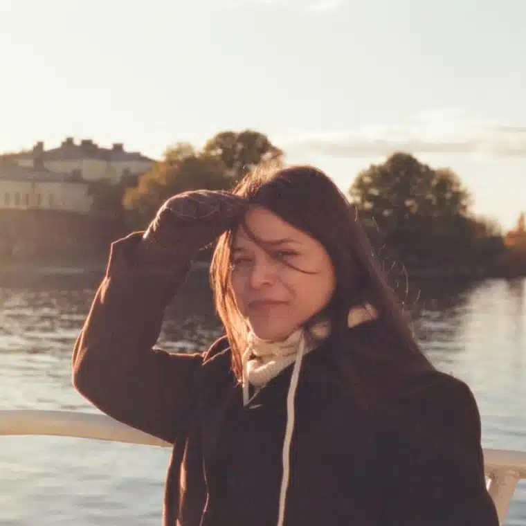 photo of a woman with brown hair and a jacket and gloves in front of a lake putting her hand up in front of her face