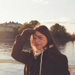 Substitute Portrait for Zaida photo of a woman with brown hair and a jacket and gloves in front of a lake putting her hand up in front of her face